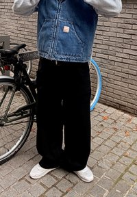 Denimvest med frontlomme, grå langærmet skjorte, løstsiddende sorte bukser og hvide sneakers. Cykler synlige i baggrunden.