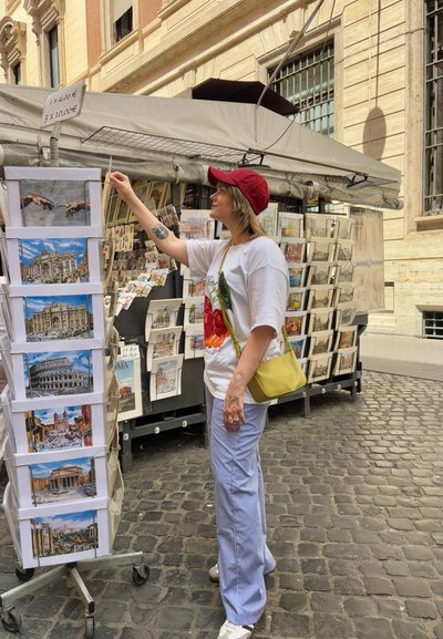 Un expositor de postales con diversas impresiones artísticas, que muestra a una mujer con un gorro rojo, camisa blanca y pantalones a rayas, extendiendo la mano para alcanzar una postal. Bolsa amarilla.