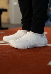 Biele nazúvacie topánky s hladkým, textúrovaným povrchom a jemnými vlnkovými vzormi; v páre s čiernymi teplákmi na červenej podlahe.