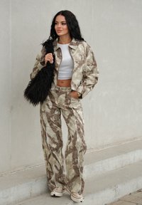 Beige och brun sammetsjacka med camouflagemönster och vida byxor, tillsammans med en vit crop top och en svart fluffig väska, allt i kombination med vita sneakers.