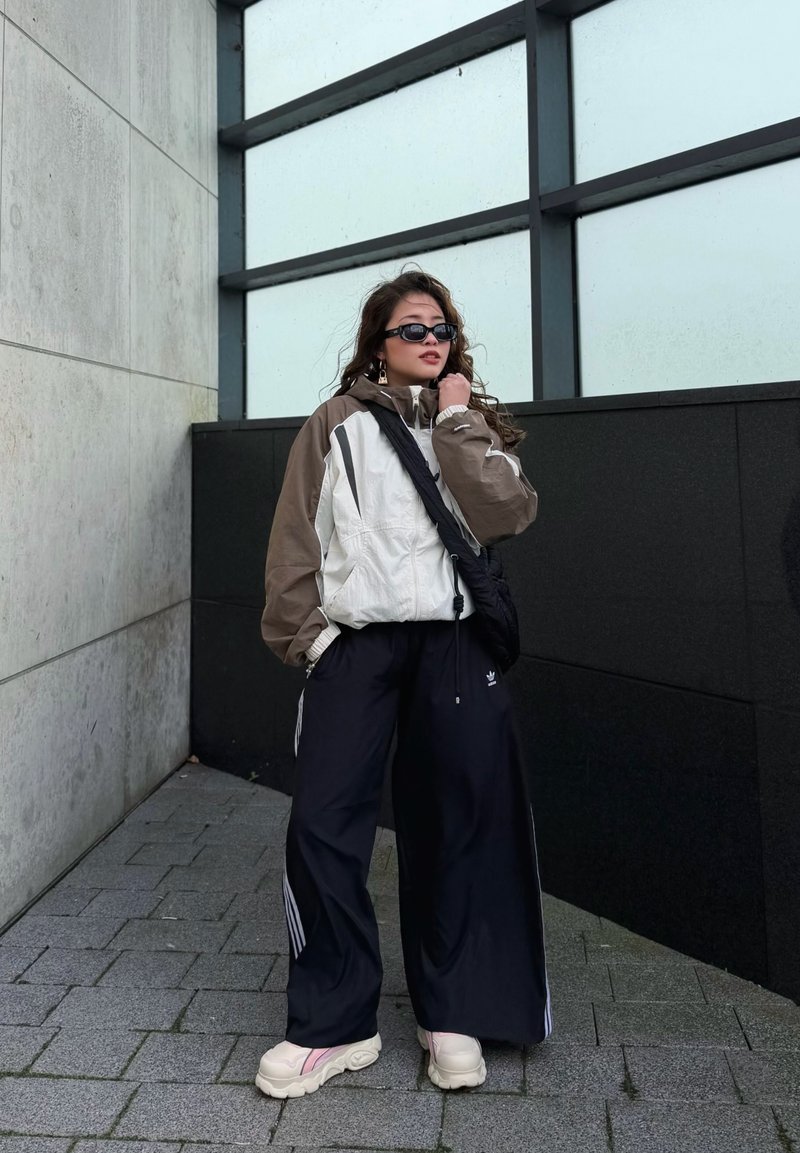 Jeune personne portant des lunettes de soleil, veste beige et blanche, pantalons larges noirs, baskets blanches, debout sur un trottoir gris près d’un bâtiment moderne.