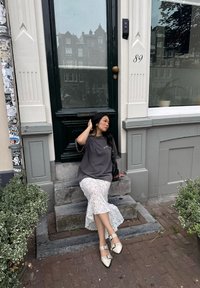 Grijze oversized t-shirt, witte kant mid-length rok, beige puntige ballerina's met studaccenten, zittend op stenen trappen bij een groene deur.