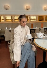 Fehér texturált blézer krémszínű garbó felett, világoskék farmerrel párosítva. Fehér csészét tart egy csészétartón, mellette fehér teáskanna áll.