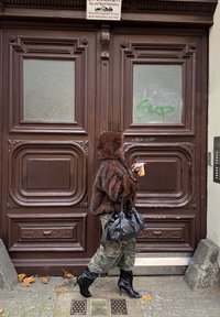 Personne portant un manteau en fourrure marron à capuche, un pantalon camouflage, des bottes noires, et portant un sac noir, marchant devant une grande porte ornée marron.
