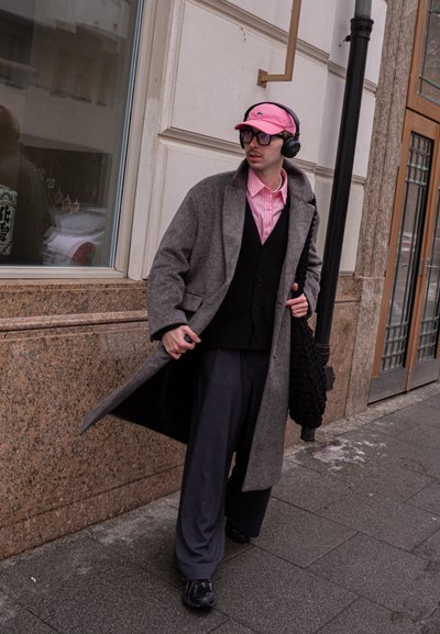 Joven con abrigo gris, gorra rosa, auriculares y gafas caminando por la acera de la ciudad llevando una bolsa negra con textura.