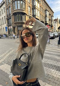 Jovem mulher com uma blusa cinza oversized e grandes óculos de sol segura uma bolsa preta, posando numa rua de calçada de cidade com edifícios históricos.