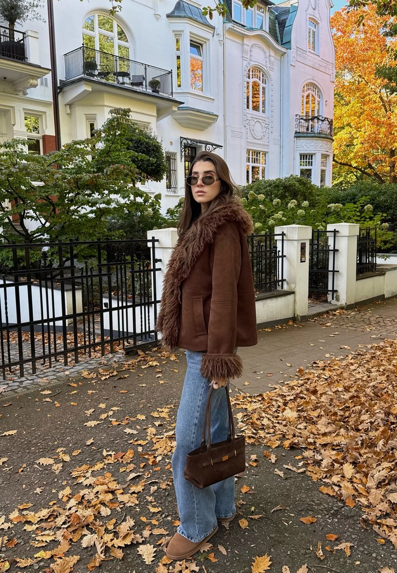 Chaqueta de piel sintética marrón con capucha, combinada con jeans acampanados de color azul claro y botas de tobillo marrones. Lleva un pequeño bolso marrón. Hojas de otoño en el suelo.