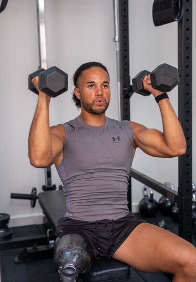 Mancuernas de forma hexagonal, con recubrimiento de goma negra. Pesas individuales, con una camiseta sin mangas gris y pantalones cortos negros.