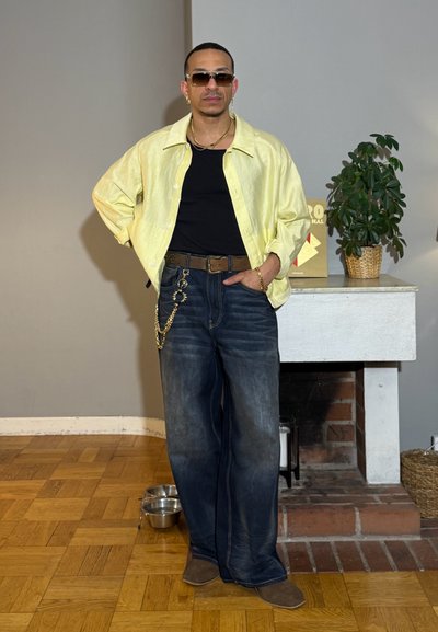 Hombre con gafas de sol, camisa amarilla sobre top negro, vaqueros anchos con cadena, de pie en el interior cerca de la chimenea con planta en la repisa.