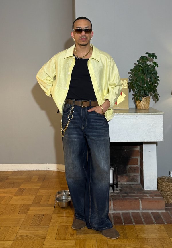 Mężczyzna w okularach przeciwsłonecznych, żółta koszula na czarnej bluzce, szerokie jeansy z łańcuchem, stoi w pomieszczeniu obok kominka z rośliną na półce.