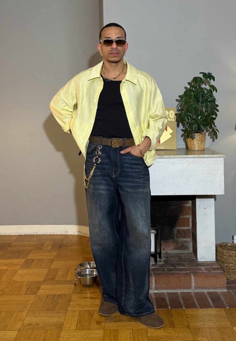 Homme portant des lunettes de soleil, chemise jaune sur un top noir, jean large avec chaîne, debout à l'intérieur près de la cheminée avec une plante sur la tablette de la cheminée.