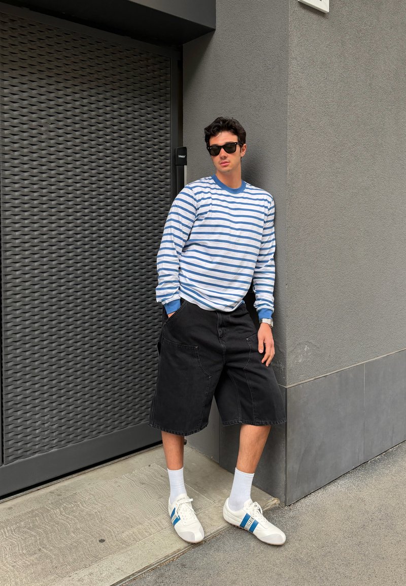 Jeune homme portant des lunettes de soleil noires, une chemise à manches longues rayée bleu et blanc, un short noir, des chaussettes blanches et des baskets blanches, appuyé contre un mur gris.