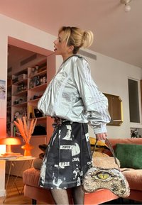 Veste argentée et réfléchissante avec des manches rayées, associée à une jupe noire imprimée avec du texte. Tient un sac à main texturé unique avec un motif d'œil.