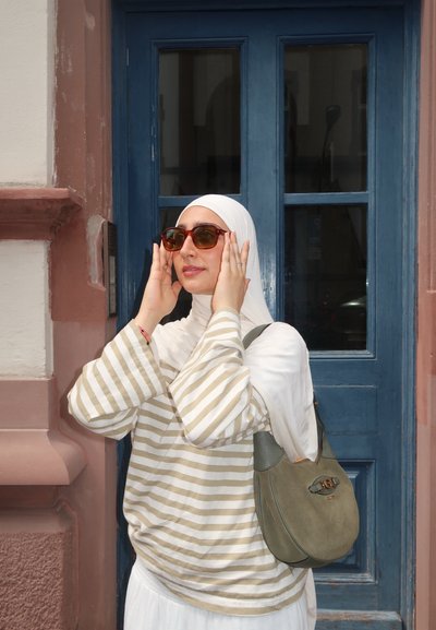 Top de gran tamaño a rayas beige y blancas con un hijab blanco, gafas de sol marrones y un bolso de ante verde, frente a un fondo de puerta azul.