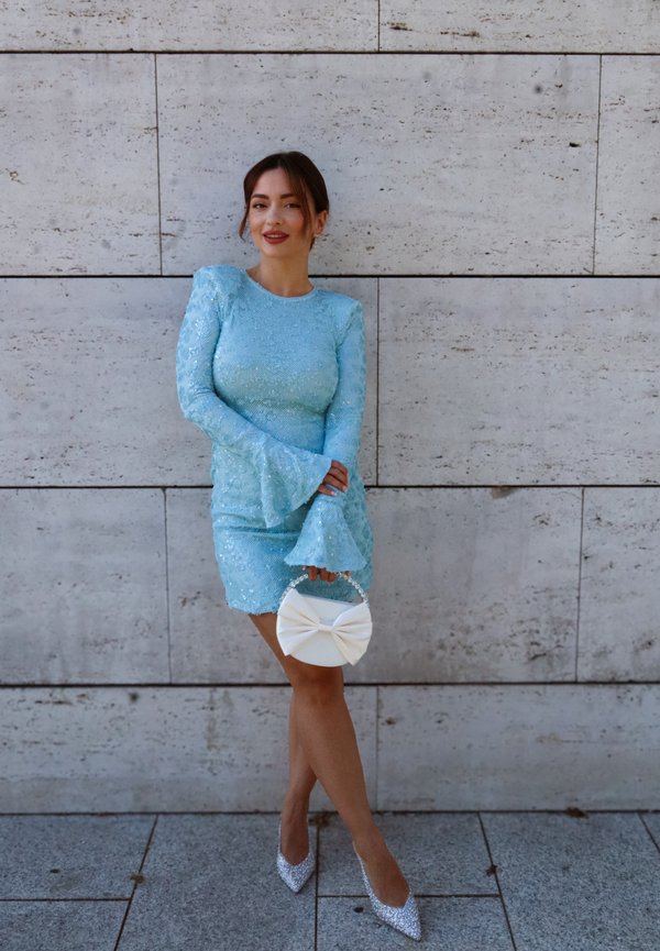 Femme portant une robe bleu clair à manches longues et des talons argentés, tenant un sac à main blanc avec un grand nœud, debout contre un mur carrelé.