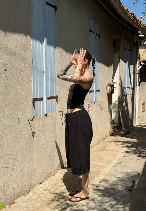 Tattooet kvinde i sort tanktop og shorts står barfodet i en solbelyst gyde og justerer håret med hænderne nær blå skodder.