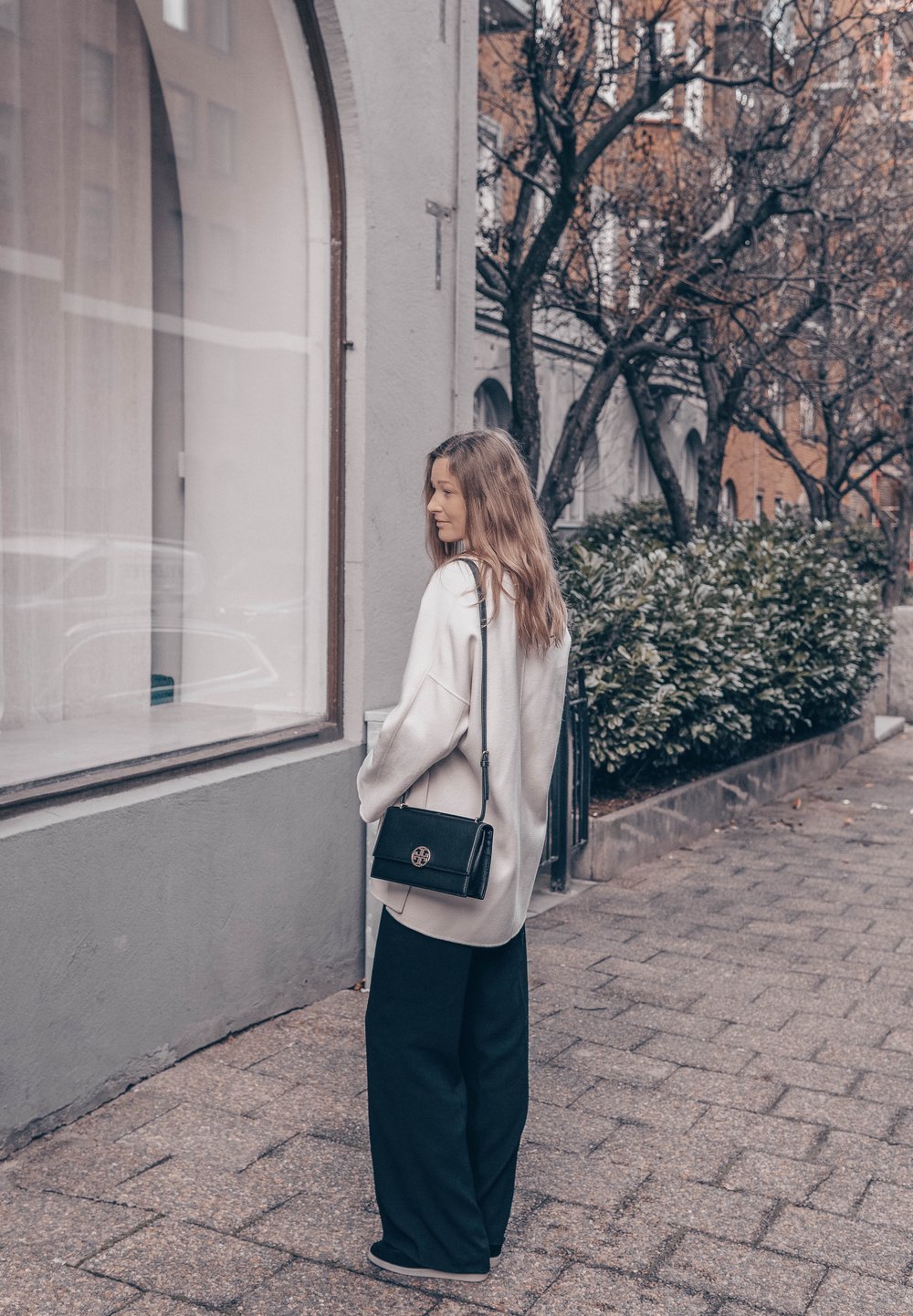 Preveliki bež pulover, crne široke hlače i mala crna torba preko ramena s logom. Teksturirana trotuarska površina i urbani pejzaž.