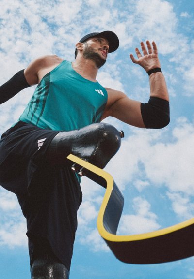 Atleta con una camiseta sin mangas deportiva color teal y pantalones cortos negros, utilizando una prótesis de pierna con una cuchilla de carrera amarilla y negra, contra un fondo de cielo azul.