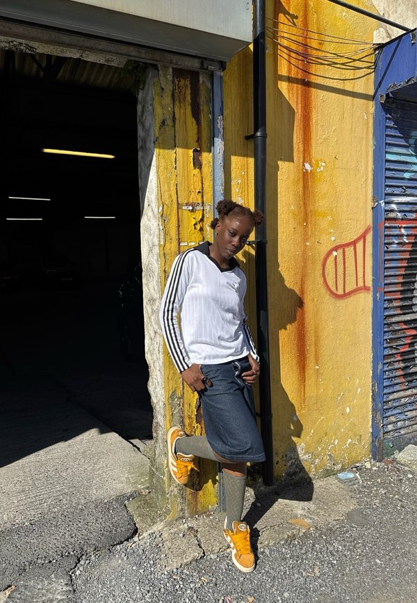 Jeune personne avec deux chignons portant un t-shirt blanc à manches longues et des baskets orange, appuyée contre un mur jaune avec des graffitis, sous le soleil.