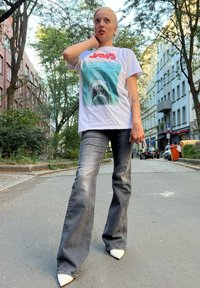 Weißes Grafikt-Shirt mit einem Hai-Motiv und rotem "Jaws"-Text. Kombiniert mit ausgestellten grauen Jeans und weißen spitzen High Heels. Urbaner Hintergrund.