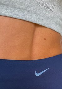 Haut gris court avec le logo "Casall" au-dessus d'une ceinture bleu marine ornée d'un swoosh Nike bleu clair ; texture de peau lisse visible.