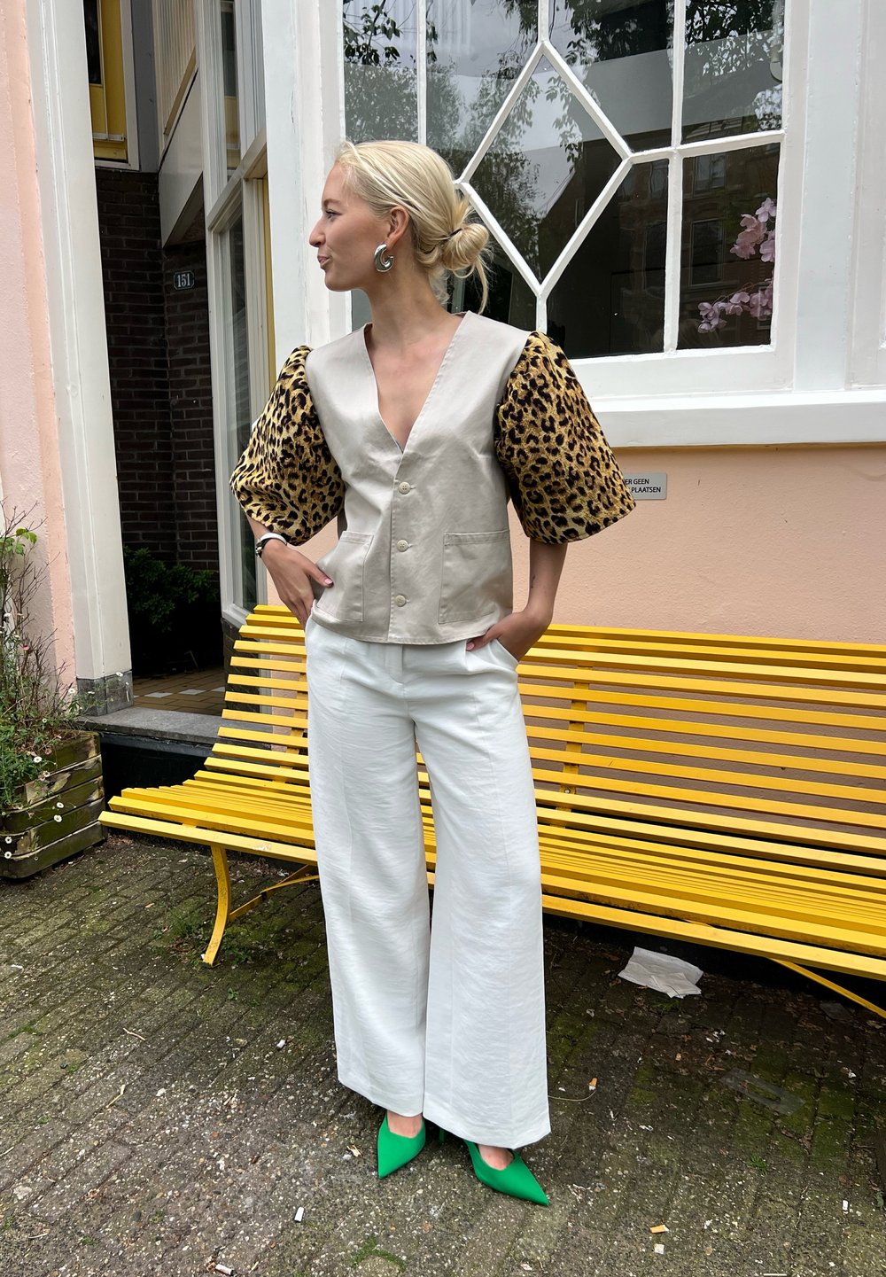 Bežinė trumpa liemenė su leopardų rašto pūstomis rankovėmis, derinama su plačiomis baltomis kelnėmis ir žaliais smaigalio formos aukštakulniais, ant gelsvo suolo.