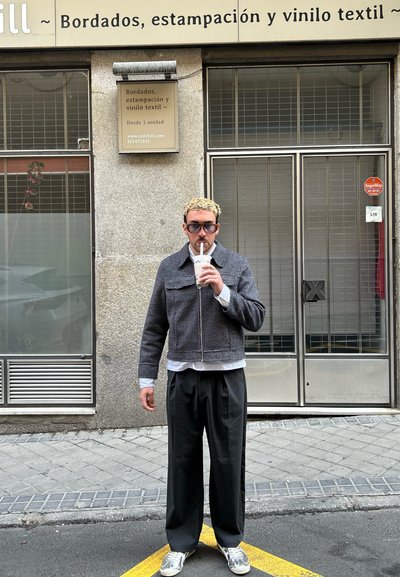 Chaqueta gris con estampado y cremallera, camisa blanca debajo, pantalones anchos negros y zapatillas grises. Sosteniendo una bebida con una pajita.