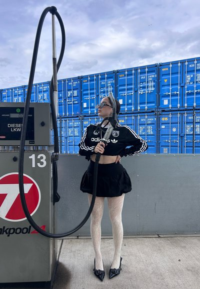 Chaqueta cropped negra de Adidas, falda negra, medias blancas con estampado y tacones negros. De pie junto a una bomba de combustible con mangueras, contenedores azules al fondo.