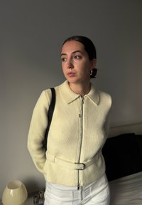 Pulover texturat de culoare crem, cu guler, fermoar frontal și detaliu cu curea. Purtați-l peste pantaloni albi, cu o geantă cu bretele negre.