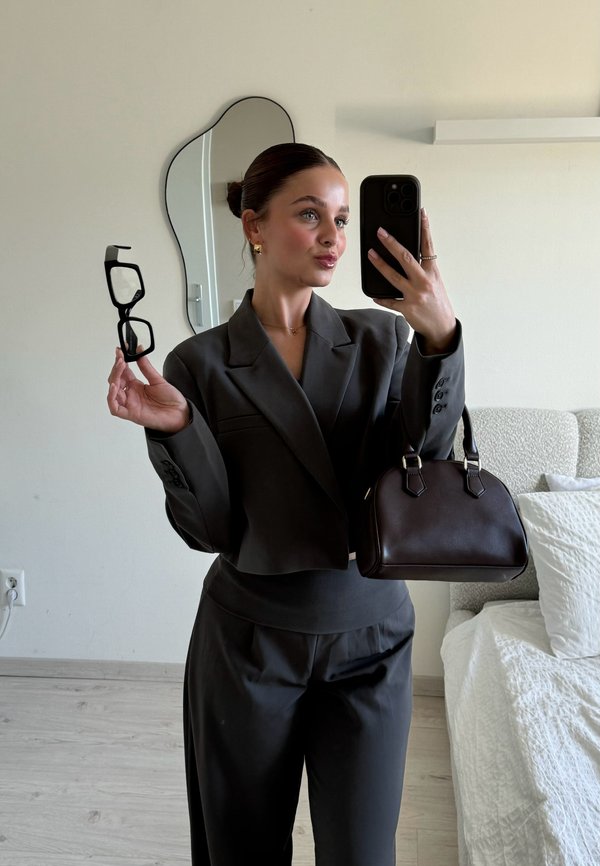 Mørkegrå cropped blazer med store klapper, matchende bukser, holder sorte briller og en lille mørkebrun taske. Neutral baggrund.