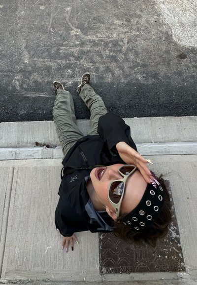 Persona con gafas de sol y cinta negra en la cabeza, recostada parcialmente sobre la acera y la calle, mirando hacia arriba con la mano en la frente.