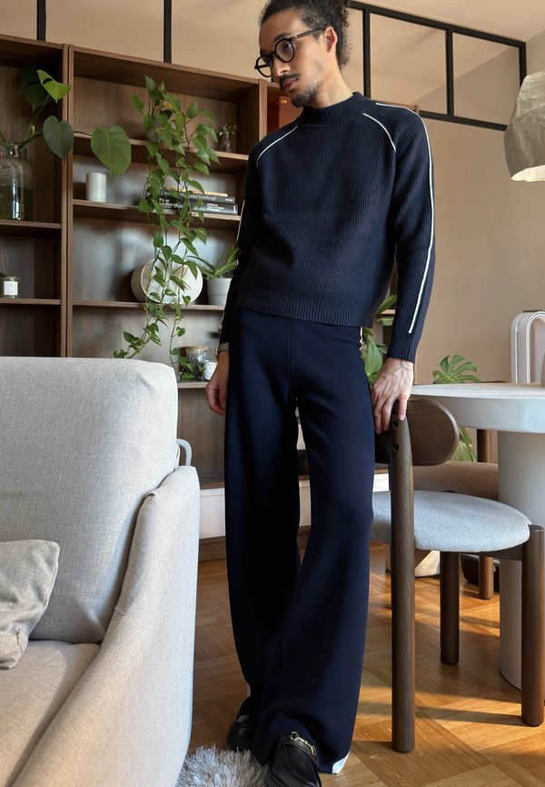 Mann iført svart genser og vide bukser, står og lener seg på en stol i en moderne stue med planter og møbler i tre.