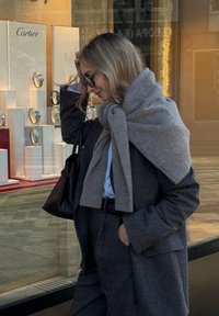 Grå uldfrakke, gråt strikket tørklæde viklet om skuldrene, sort håndtaske, st�ående foran en smykkebutik med ure i udstillingsvinduet.