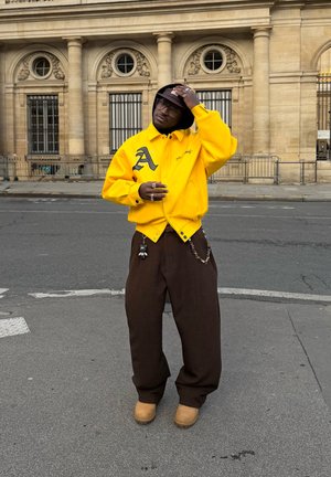 Persona con chaqueta amarilla y pantalones marrones ajustándose un sombrero tipo pescador negro en una calle de la ciudad con un edificio histórico de fondo.