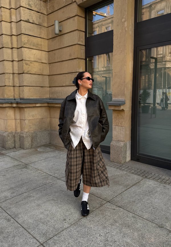 Woman wearing black leather jacket, white shirt, plaid wide-leg pants, black sneakers, and sunglasses standing near stone building with large windows.