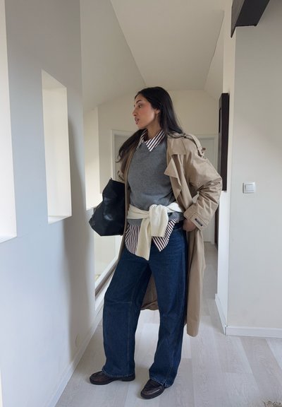 Gabardina beige sobre un suéter gris, camisa de rayas y jeans anchos azul oscuro. Bolso negro y zapatos oscuros completan el conjunto.