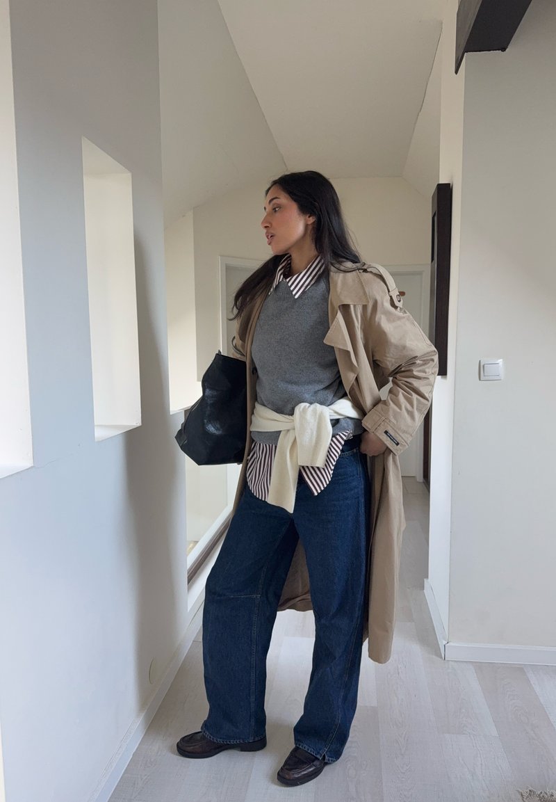 Trench-coat beige sur un pull gris, une chemise rayée et un jean large bleu foncé. Sac noir et chaussures foncées complètent le look.