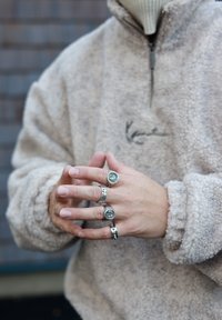 Pull polaire à fermeture éclair en beige avec tissu texturé. Les mains affichent quatre bagues en argent, avec des détails gravés et des designs variés.