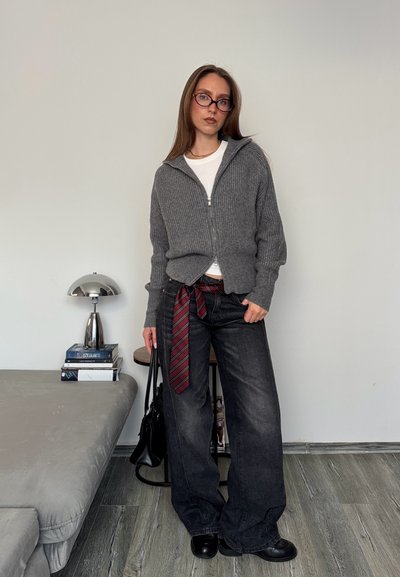 Mujer con suéter gris con cremallera, camisa blanca, pantalones anchos negros con cinturón de corbata a rayas rojas, gafas, sosteniendo un bolso negro, de pie en una habitación moderna.
