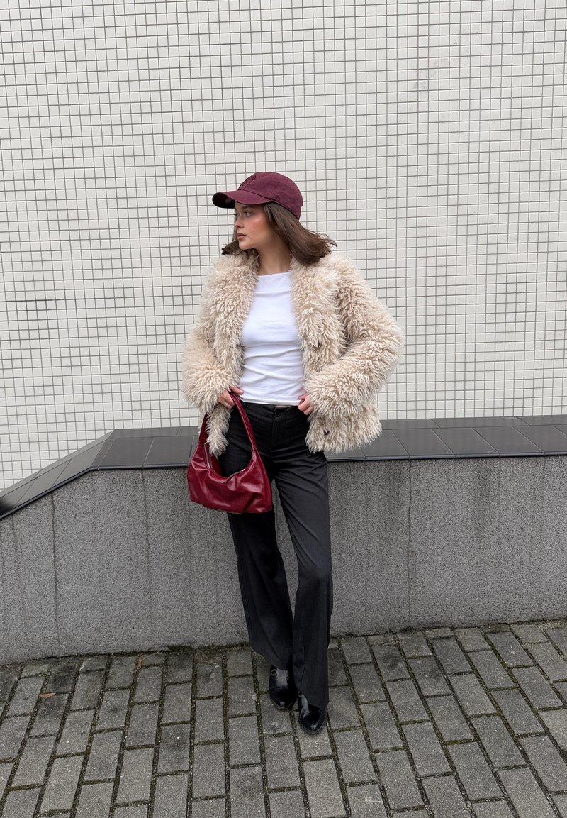 Giovane donna che indossa una giacca beige soffice, camicia bianca, pantaloni neri, scarpe nere e un berretto bordeaux, con una borsa bordeaux abbinata in mano.