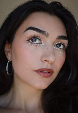 Nærbillede af et ansigt med mørkt hår, fremtrædende øjenbryn, udsmykkede øjne med sølv- detaljer og blanke brune læber. Sølv hoop-øreringe er synlige.