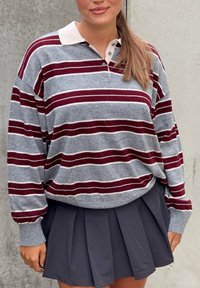 Kobieta ubrana w szarą i bordową pasiastą bluzkę polo z długim rękawem, wpuszczoną do ciemnoszarej plisowanej spódnicy, stojąca na tle betonowej ściany.