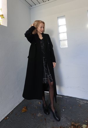 Abrigo negro cruzado de silueta larga, superpuesto a un vestido con patrón geométrico en blanco y negro, combinado con mocasines negros.