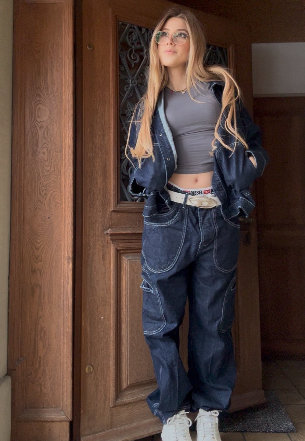 Jeune femme aux longs cheveux portant des lunettes, un top court gris, une veste en jean oversize et un pantalon, ceinture blanche, se tenant près d'une porte en bois.