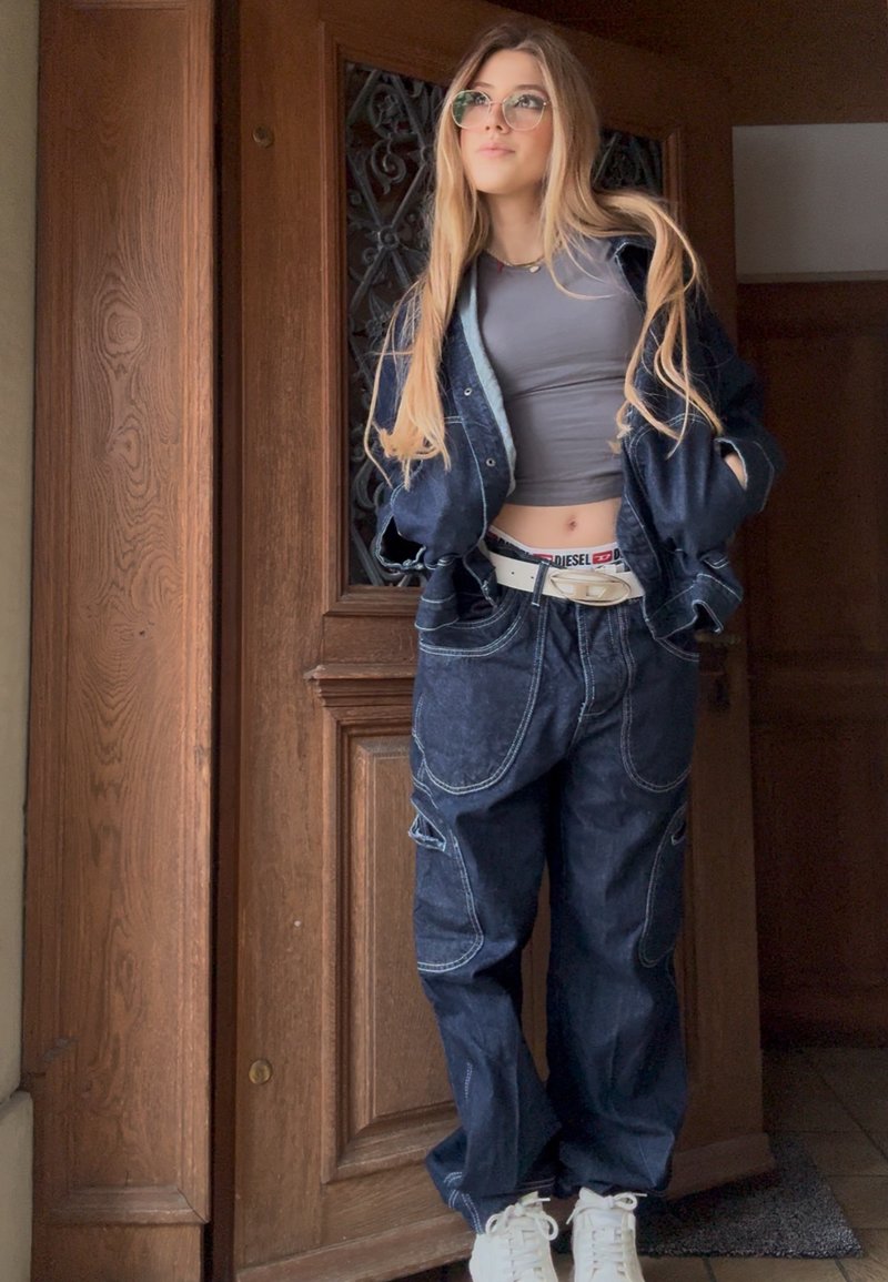 Junge Frau mit langen Haaren, die eine Brille, ein graues Croptop, eine übergroße Jeansjacke und Hose sowie einen weißen Gürtel trägt, steht neben einer Holztür.