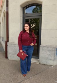 Kobieta w bordowej koszuli i niebieskich dżinsach, trzymająca czerwoną torebkę, stojąca przed budynkiem z ciemnymi drzwiami ramowymi.