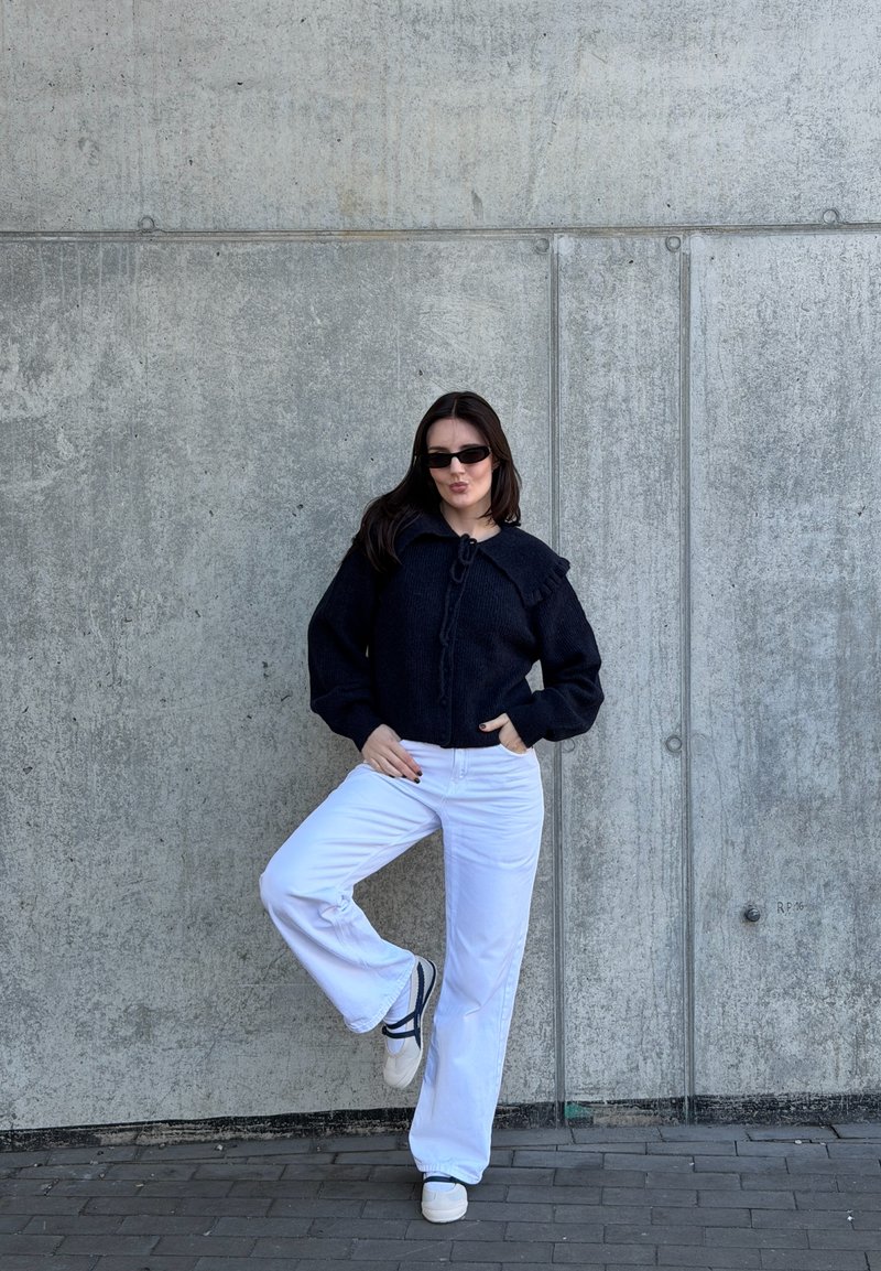 Donna con maglione nero, pantaloni bianchi a gamba larga e scarpe da ginnastica bianche posa con una gamba sollevata contro un muro di cemento indossando piccoli occhiali da sole neri.