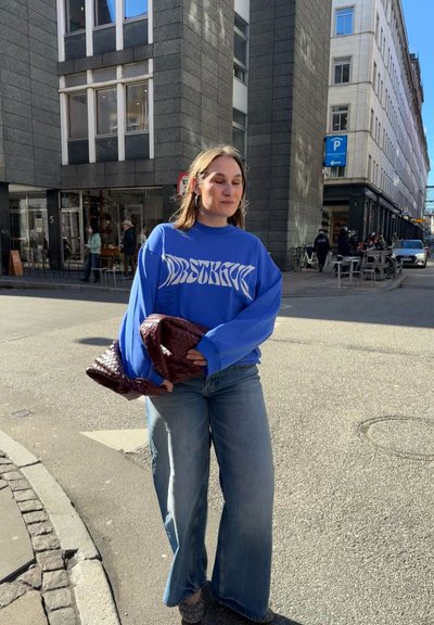 Joven mujer con sudadera azul y vaqueros anchos sostiene un gran bolso de mano marrón rojizo mientras camina por una calle soleada de la ciudad.