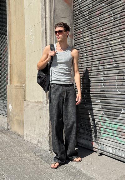 Persona vistiendo una camiseta sin mangas gris y pantalones de mezclilla negros holgados, sosteniendo una mochila negra, de pie cerca de una persiana de metal cubierta de grafitis.