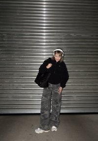 Fleece fekete dzseki, szürke álcázott, bővülő nadrágok, steppelt fekete táska és fehér tornacipő. Ezüst fejhallgató díszíti a fejét.